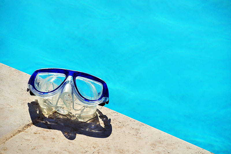 A pair of swimming goggles sit next to a brand new pool on a sunny day.